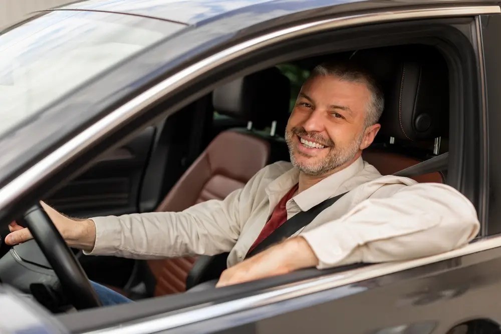Cheerful middle aged man driving auto, looking through open window and smiling at camera, happy driver enjoying car ride